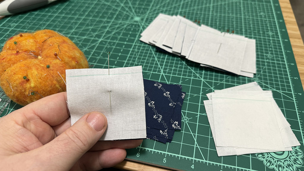 Stacks of squares, pinned together with one pin. One square held in my hand. An orange pin cushion on a green cutting mat in the background.