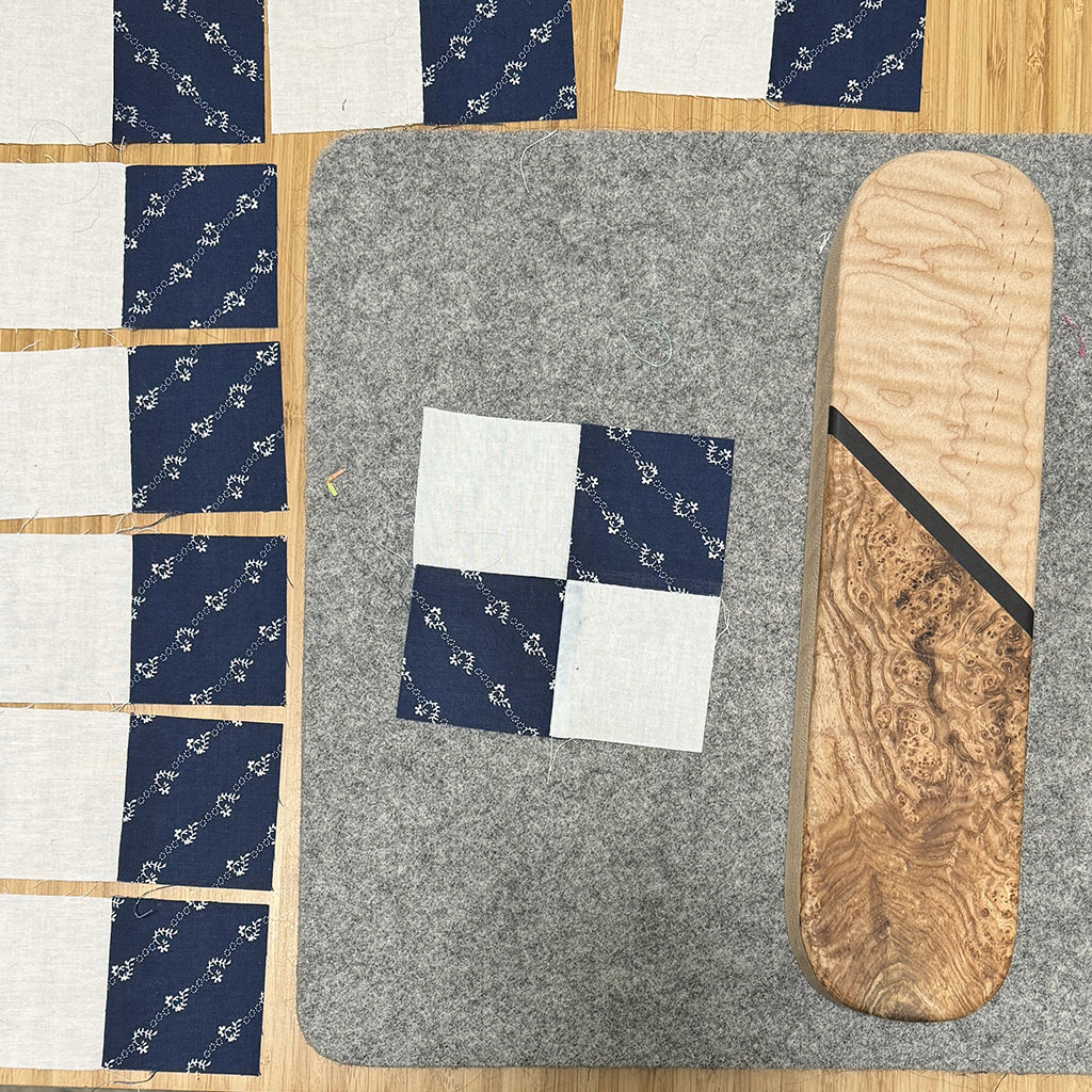 A 4-patch on a grey wool ironing mat beside a tailor clapper. A bunch of 2-patch pieces are lined up around the ironing mat.