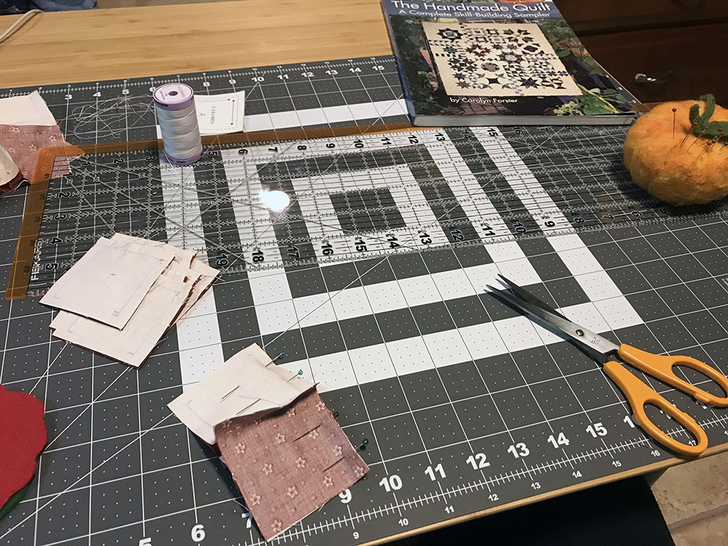 Square of fabric in red and white, pinned together. An orange pincushion is on a grey cutting mat. A pair of scissors and a large quilting ruler are also on the mat. The Handmade Quilt book is on the table.