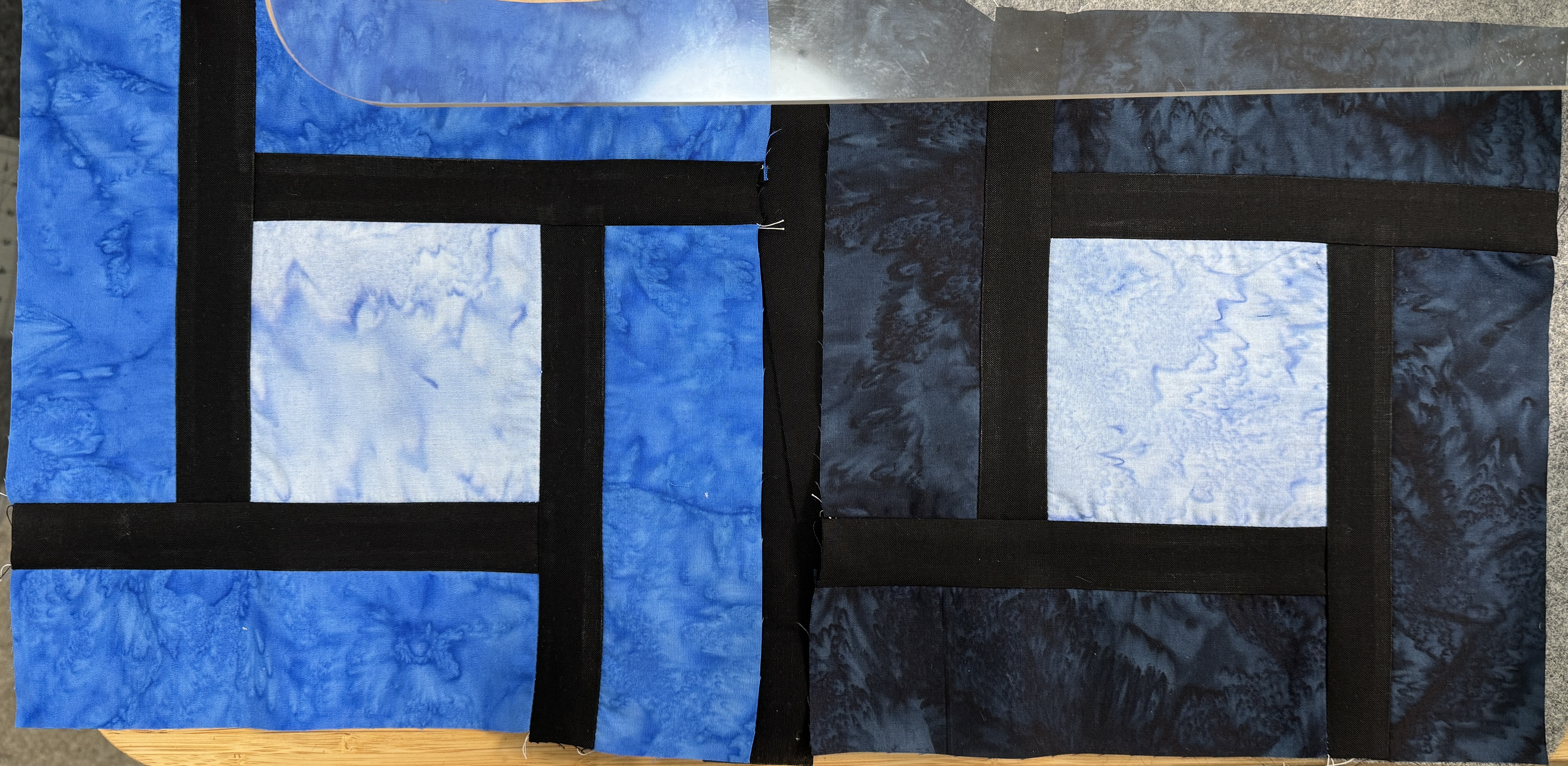 Two similar blocks of pieced fabrics. There's a light blue square in the center. Black sashing on the side of the square, extending in four directions. With blue fabric to finish a 10 inch block.