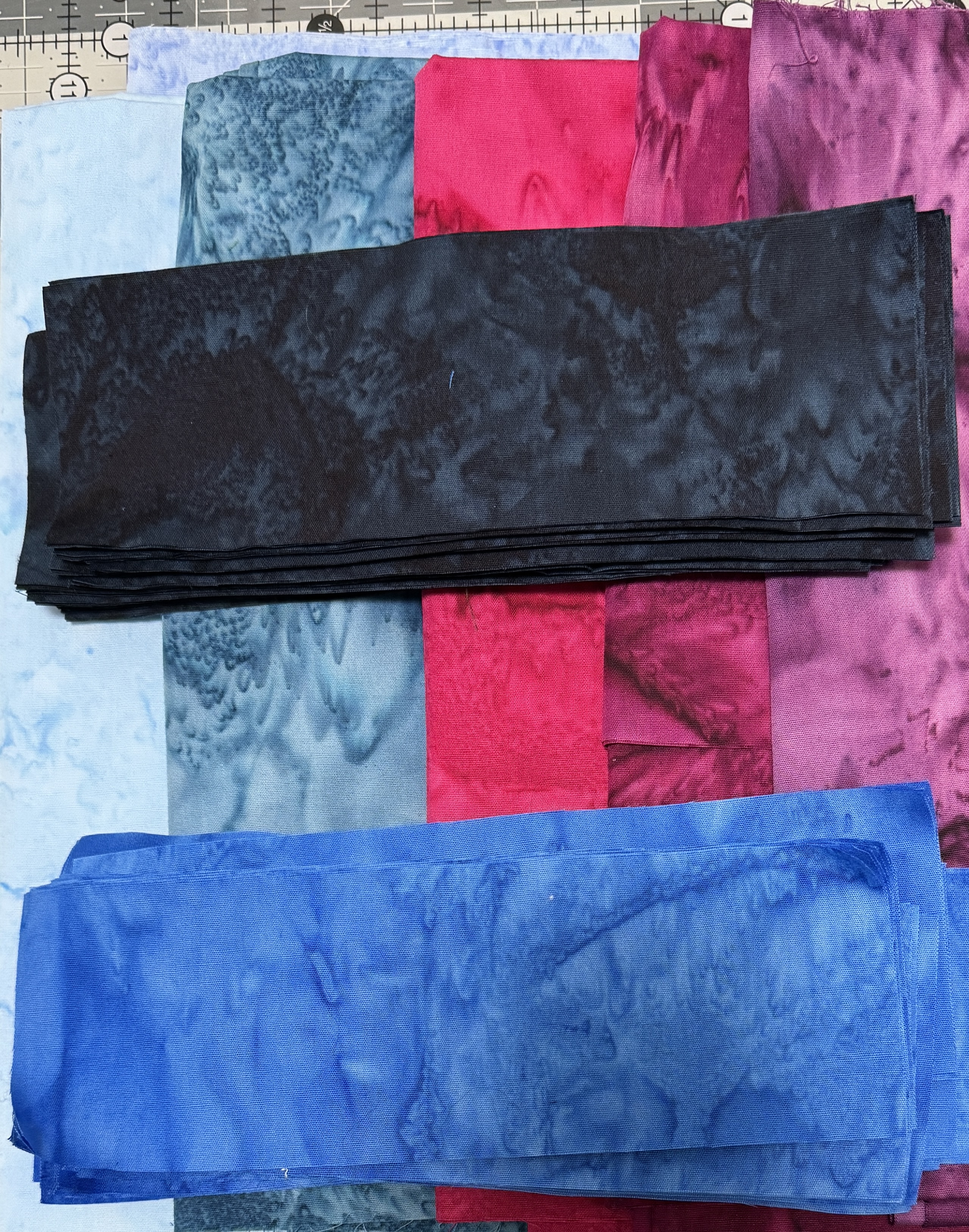 A stack of dark blue batik strips and a stack of medium blue batik strips over several other batiks in blues, reds, and purples.