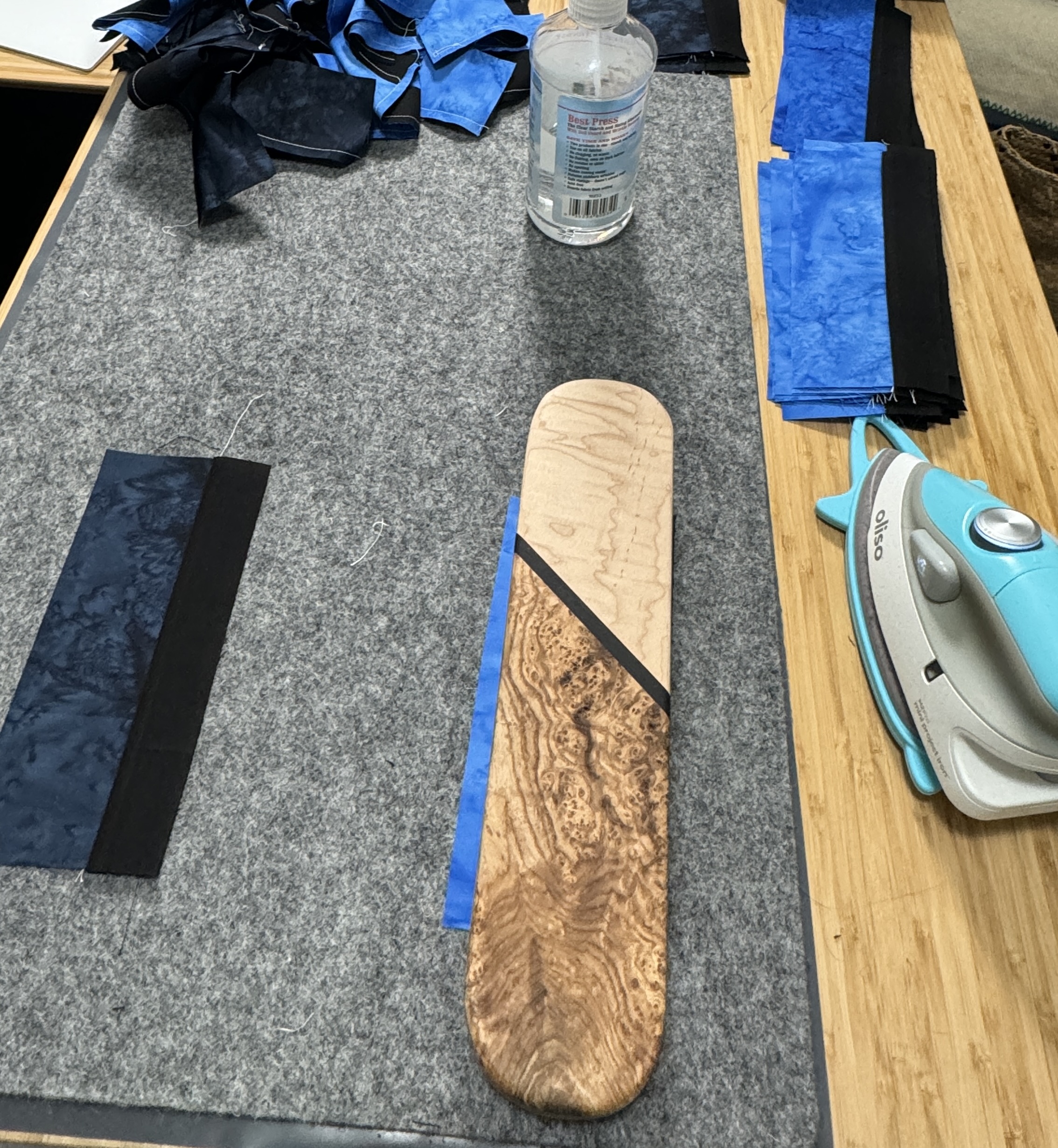 A 3 inch x 12 inch block of hardwood on fabric, on top of a grey wool ironing mat, with a small iron beside the mat, and a bottle of starch alternative.