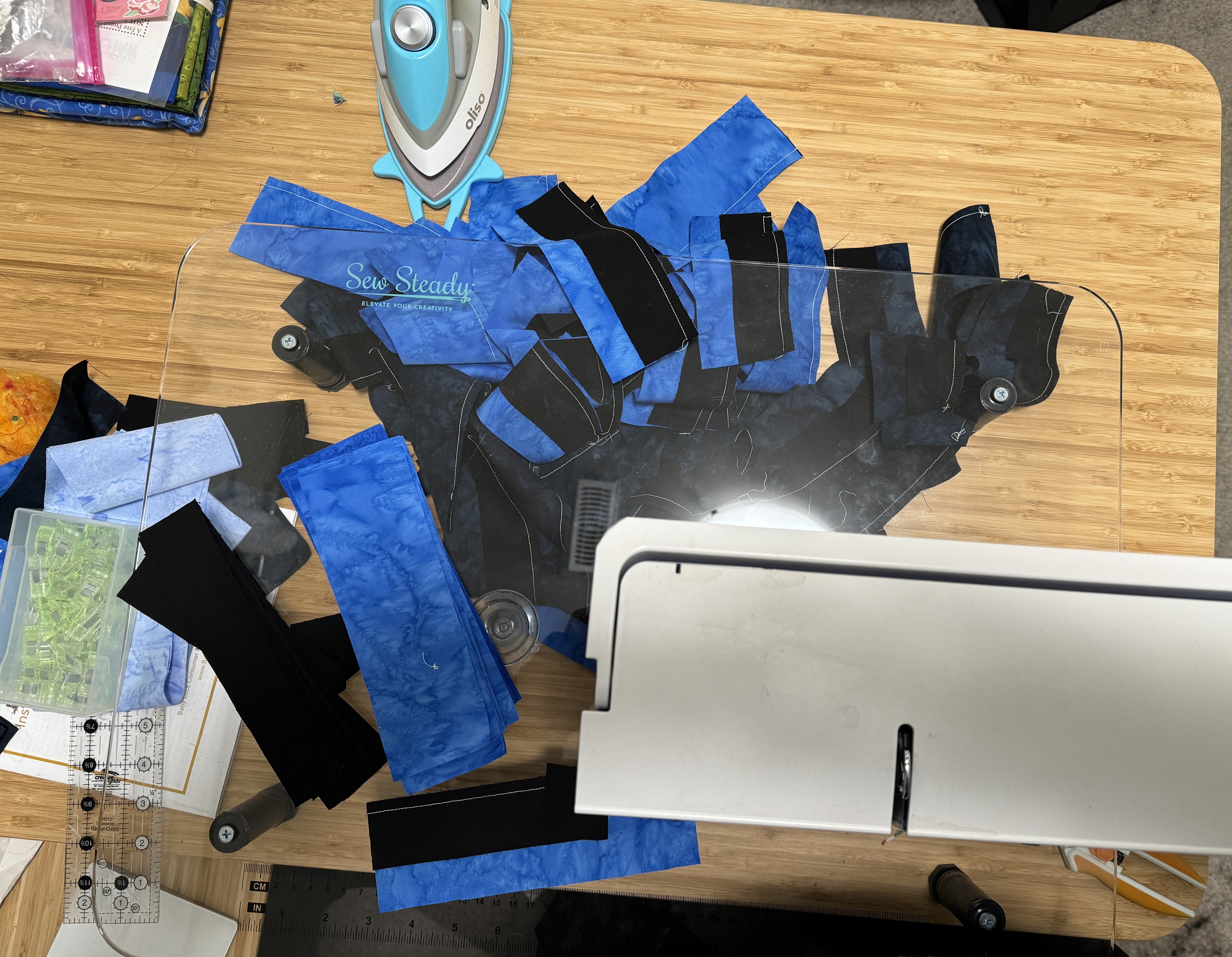 A haphazard stack of pieces of pieced fabric on a desk, under a clear sewing table extension.