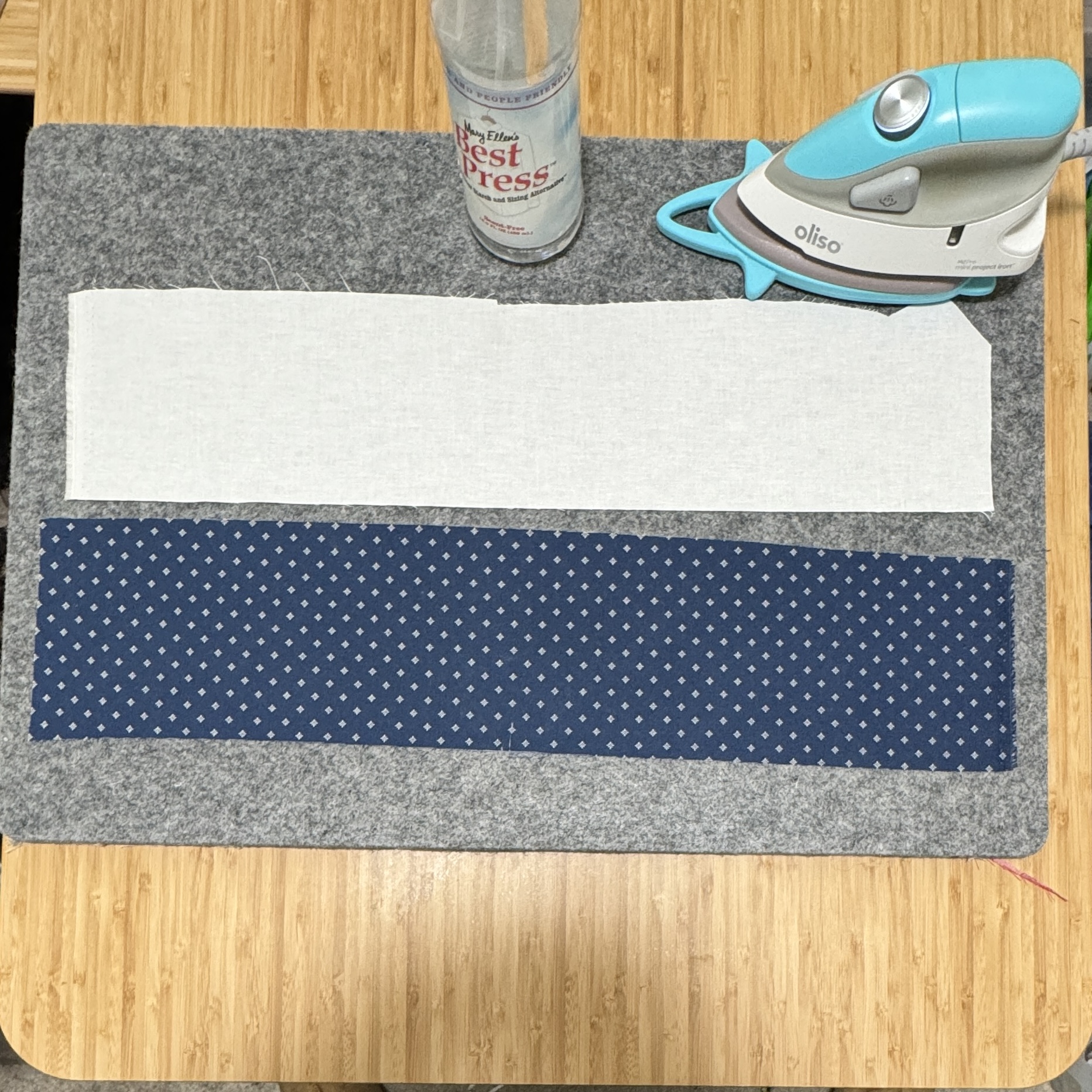 Two 4 inches by 22 inches strips of fabrics on a wool pressing mat. A small iron and a spray bottle of Best Press beside the fabric.