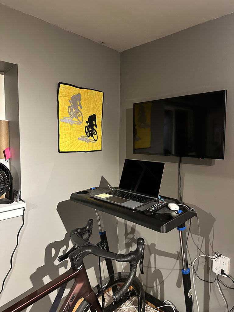The cyclist quilt hanging on a wall beside a big monitor, and a bicycle on a track. A laptop is in front of the bike