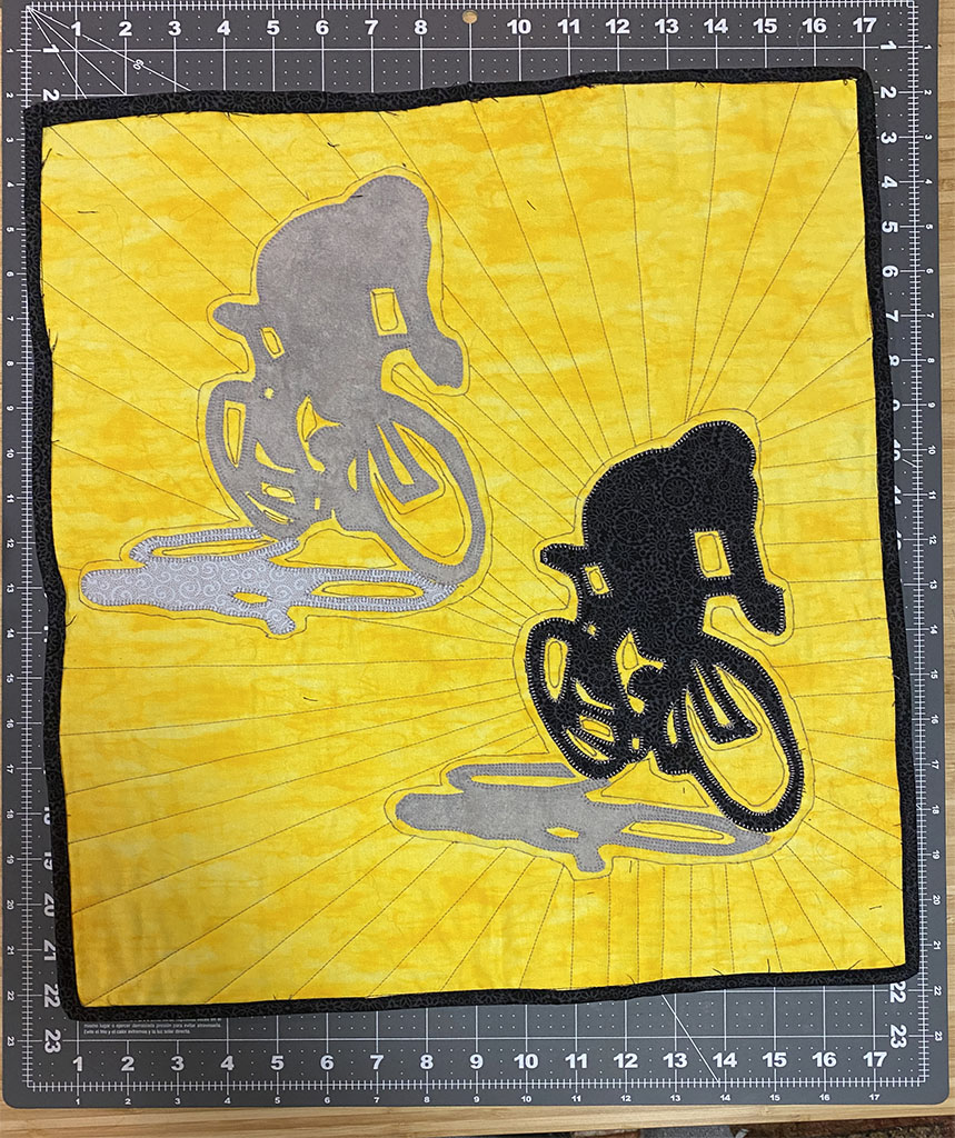 17 inches by 21 inches quilt. The background is yellow. There are two silhouettes of cyclists on racing bikes. The one in the bottom right is nearly black. The one in the top left is grey. They each have a lighter grey shadow beside them. The binding is black. The quilting is basically a bunch of straight lines coming from the cyclists and going to the edges of the quilt