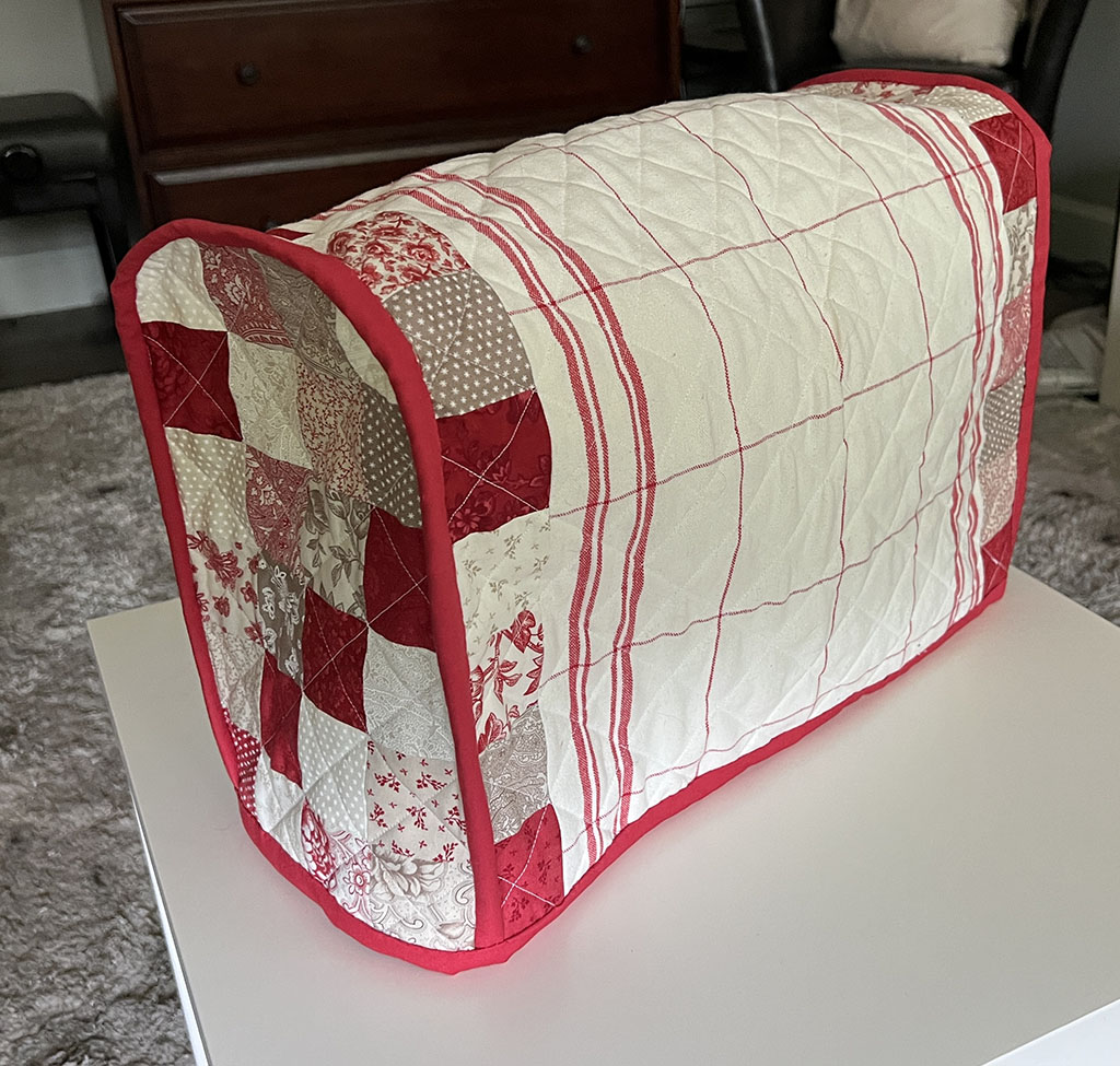 Sewing machine cover in reds and whites. The side and edges are a series of 2 and a half inch squares. The body of the cover is white with red vertical stripes