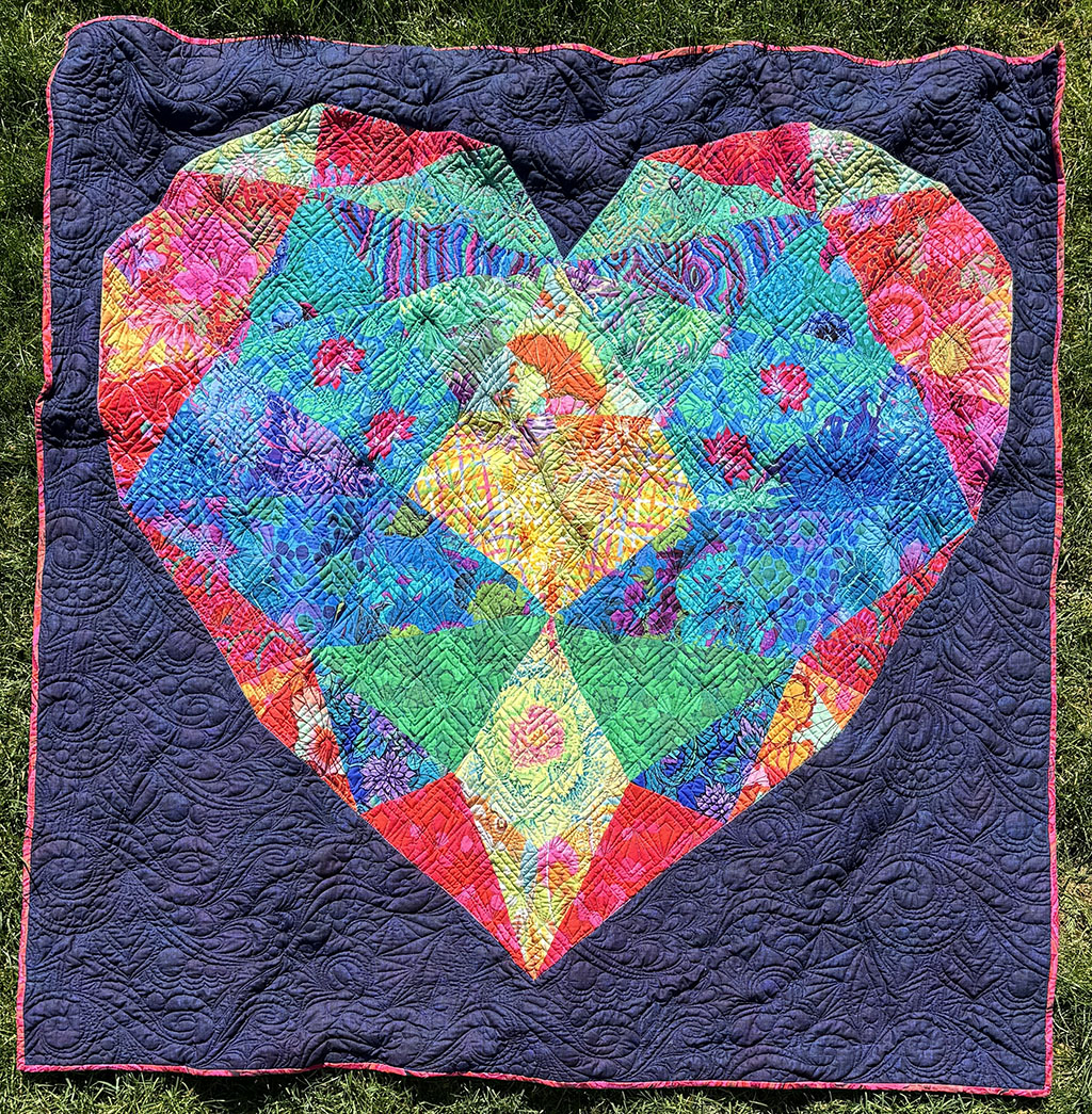 60 inch square quilt on the lawn. There is a large heart made of red, blue, green, yellow, and orange prints. The outside of the heart is dark purple.