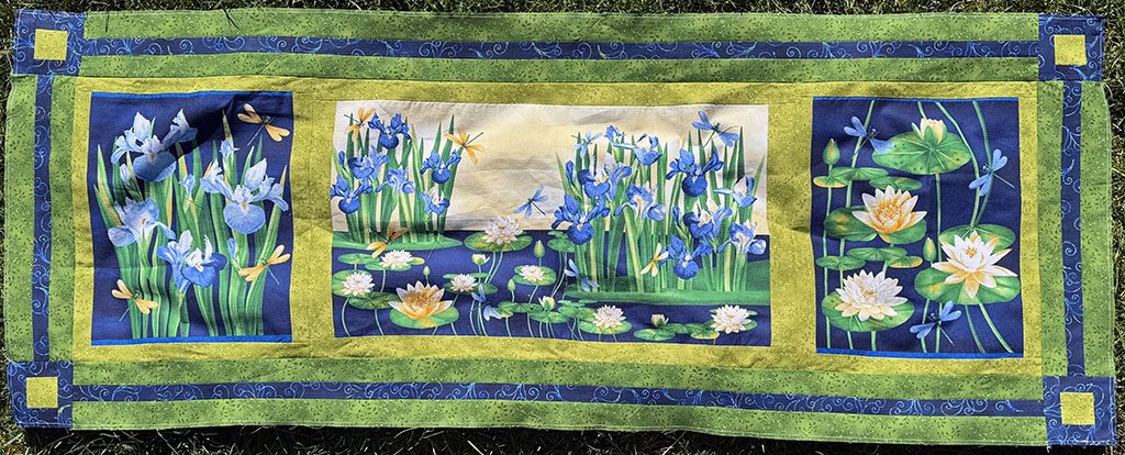 53 inches by 20 inches large piece of pieced fabric with 3 panels and borders. Colors are all in blues, greens, and yellows. The panels have irises and dragonflies