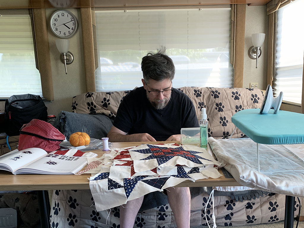 Nic behind a table covered with patchwork blocks, an open book, a small ironing board with an iron. He is focused on his hand sewing, unaware of the photo being taken