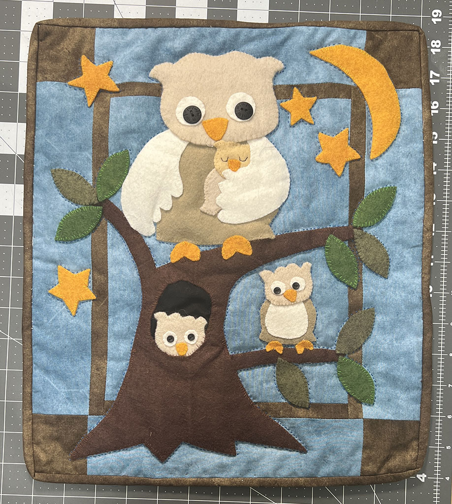 A 13 inches by 16 inches wall hanging quilt. It is all felt applique of a mama owl on top of a tree, holding a sleeping tiny owl. In the tree hole is a young owl. On another branch is another little owl. There are orange stars and crescent moon..