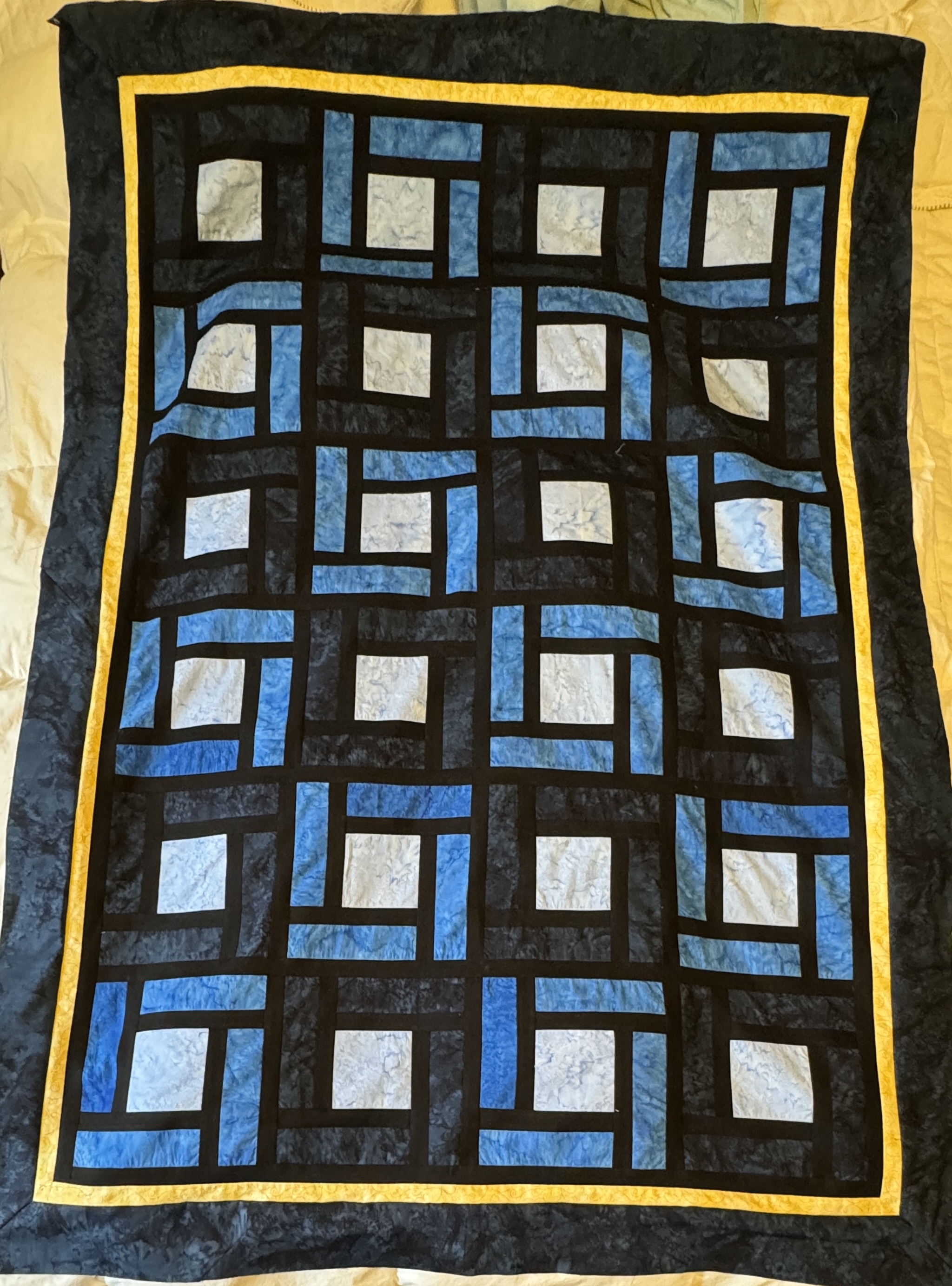 A 44 inches by 64 inches quilt top. 6 rows of 4 blocks. Each block has a center square, with black sashing and medium or dark blue around it. There is a 1 inch black sashing around the whole thing, and a 1 inch yellow sashing around that, and then a 4 inches dark blue batik border to finish it all.