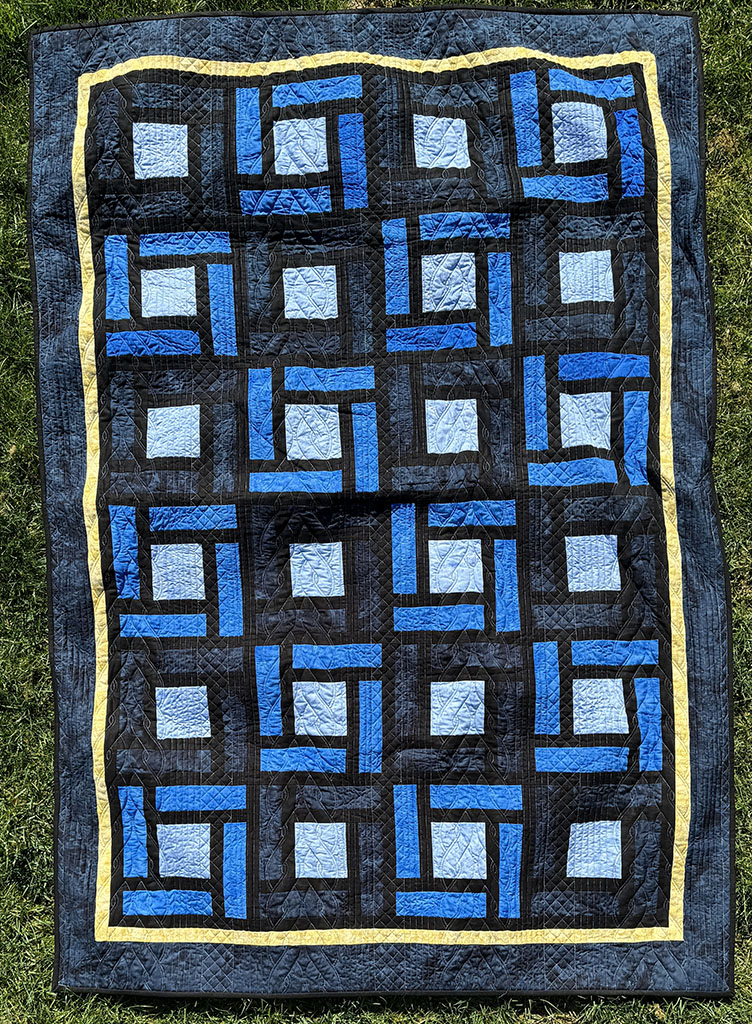 Wrap around quilt laying on the grass. It is a series of light, medium, and dark blue squares separated by black sashing. There is a yellow border on the outside, and a dark blue border around that..