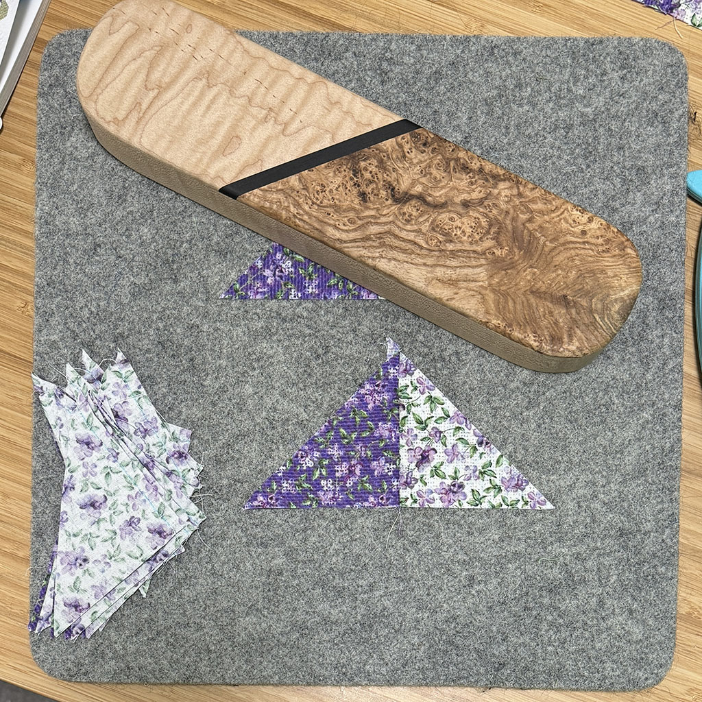 Light and dark triangle pieced with the short sides - which is the correct side. All on a grey wool pressing mat. One pieced pair is pressed open. One pair is under a clapper. A stack of unpressed pairs is on the left of the mat.