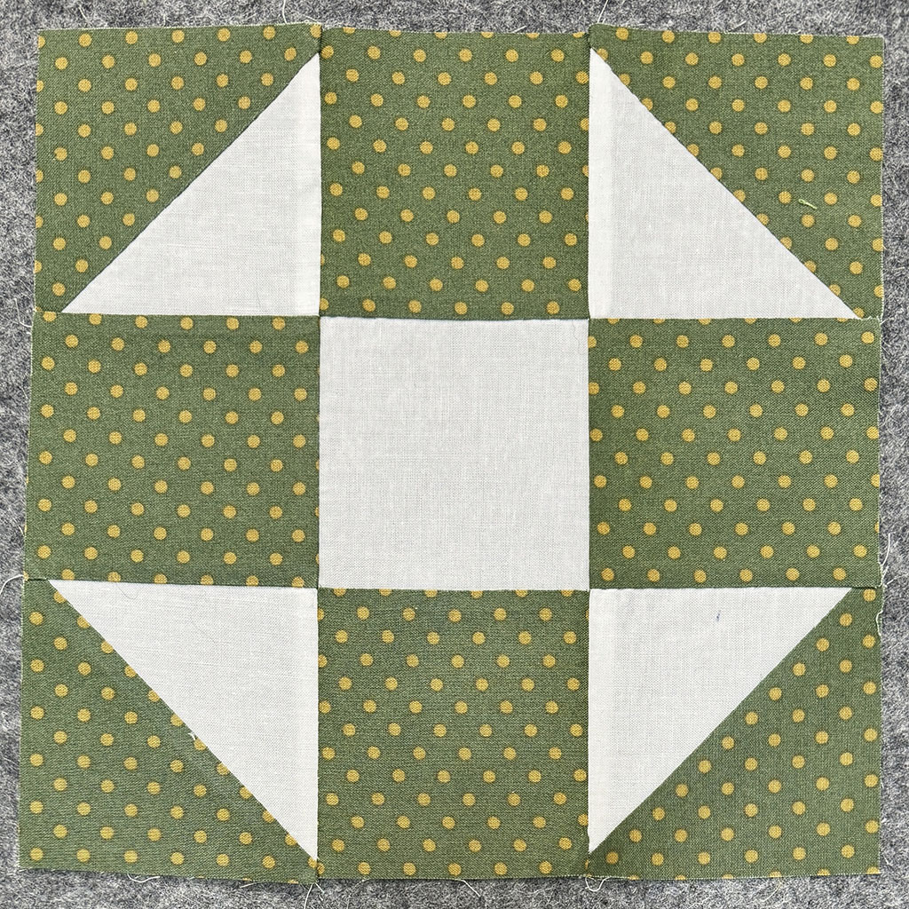 A pieced block of fabric made from off white fabric and moss green with gold polka dots fabric. Three rows of three columns. The corner squares are made of two half triangles.