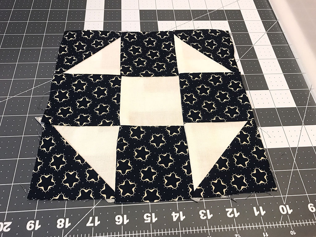 A pieced block of fabric made from off white fabric and navy blue with light white print fabric. Three rows of three columns. The corner squares are made of two half triangles.